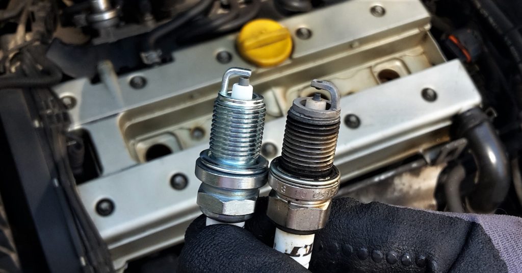 Two spark plugs with the car engine in the background. One spark plug is new, the other one is old. 