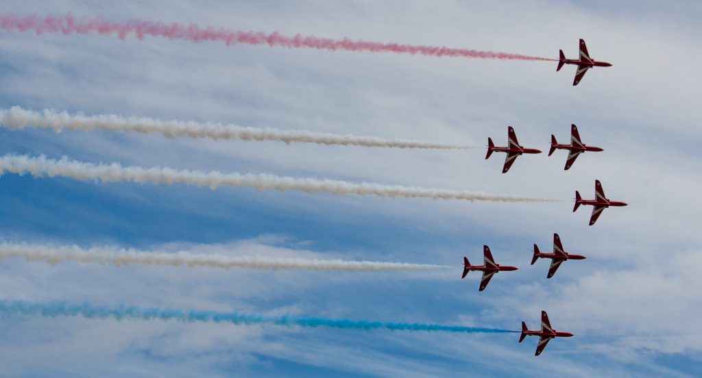 Seven aerobatic airplanes flying in a formation and leaving trails of red, white and blue smoke.