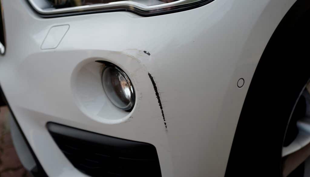A deep bumper scratch on a white, used car. The black plastic primer is exposed.